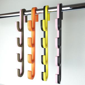 Può includere: Una fila di ganci colorati a forma di cactus appesi a una barra cromata. I ganci sono in tonalità di rosa, marrone, arancione, giallo e nero. Ogni gancio ha tre punte curve per appendere oggetti. Lo sfondo è una parete bianca.