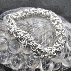 May include: A silver-colored chainmail bracelet with a woven design. The bracelet is circular and rests on a bed of clear marbles. The links are tightly interlocked, creating a textured surface. The bracelet appears to be made of metal.
