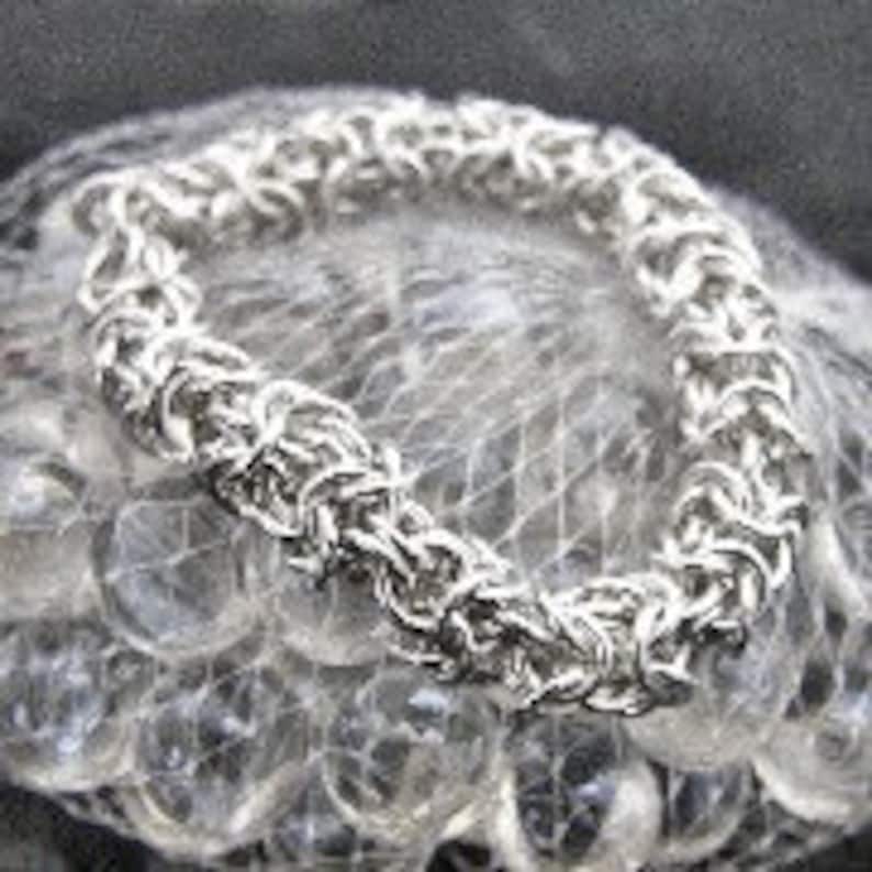 May include: A silver-colored chainmail bracelet with a woven design. The bracelet is circular and rests on a bed of clear marbles. The links are tightly interlocked, creating a textured surface. The bracelet appears to be made of metal.