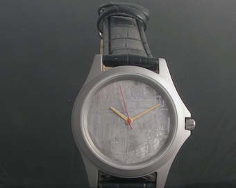 May include: A silver wristwatch with a black leather band. The watch face is made of a meteorite with a gray and white pattern. The watch has red and gold hands.