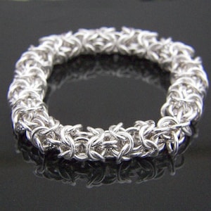May include: A silver-colored bracelet with a Byzantine chainmail design. The bracelet is circular and made of interlocking metal rings. The jewelry is displayed on a reflective black surface.