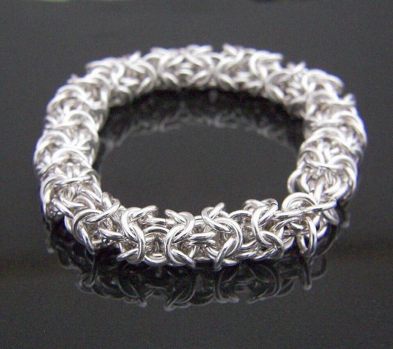 May include: A silver-colored bracelet with a Byzantine chainmail design. The bracelet is circular and made of interlocking metal rings. The jewelry is displayed on a reflective black surface.