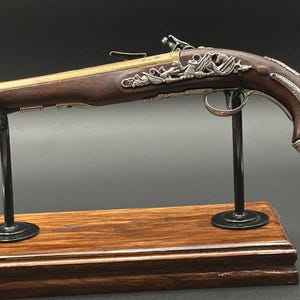 Op de afbeelding: Een replica van een vuursteenpistool met een donkerbruine houten kolf, een koperen loop en gedetailleerde zilverkleurige metalen filigraanversieringen langs de zijkant en de greep. Het wapen staat op een zwarte metalen standaard met een houten voet.