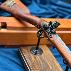Può includere: Un fucile a pietra focaia in stile antico, con un calcio in legno scuro e decorazioni in metallo. Il fucile è esposto su un supporto in legno, che ne mette in risalto la maestria artigianale. Lo sfondo è un tessuto blu scuro.