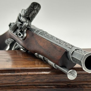 Może przedstawiać: Pistolet skałkowy z ciemnobrązową drewnianą kolbą i metalową lufą z misternymi kwiatowymi grawerunkami. Broń posiada srebrzysty mechanizm kurkowy oraz stempel umieszczony pod lufą, prezentujący szczegółowe wykończenie metalowych elementów.