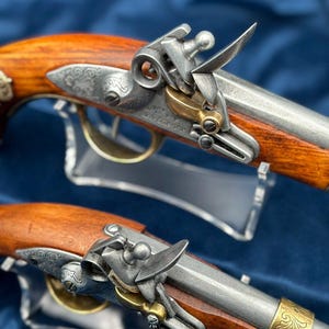 Può includere: Due antiche pistole a pietra focaia con manici in legno e dettagli metallici ornati. Le pistole presentano accenti argentati e dorati, con incisioni intricate sui componenti metallici. I manici in legno sono di un ricco e caldo colore marrone. Su sfondo blu.