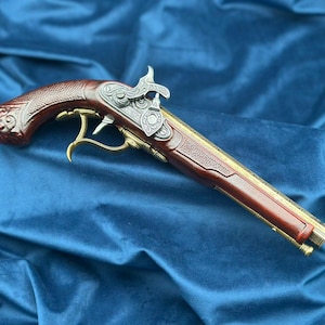 Op de afbeelding: Gedetailleerde replica van een vintage flintlock pistool. Het pistool heeft een donkerbruine houten handgreep met ingewikkelde gravures, zilverkleurige metalen accenten en goudkleurige metalen trekker- en loopdetails. Het vuurwapen wordt tegen een blauwe achtergrond getoond.