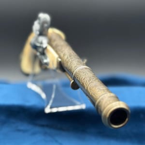 Può includere: Pistola antica in ottone con intricate incisioni floreali. La canna e l'impugnatura sono decorate con motivi ornati. La pistola è esposta su un supporto trasparente su sfondo blu, mettendo in risalto il suo design storico.