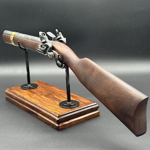 Può includere: Una pistola a pietra focaia con calcio in legno marrone scuro, canna metallica con finitura usurata e una fascia in ottone vicino alla volata. L'arma è montata su un supporto in metallo nero fissato a una base rettangolare in legno lucido.