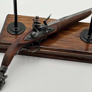 Può includere: Una replica di pistola a pietra focaia con calcio in legno marrone scuro, canna in metallo argentato e un pomolo decorato color argento a forma di testa di creatura. Il meccanismo include un cane dettagliato montato su una base in legno.