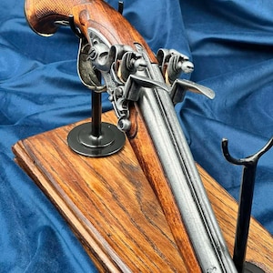 Può includere: Una pistola a pietra focaia d'epoca con manico in legno e canna in metallo, esposta su una base di legno. La pistola presenta un'intricata lavorazione del metallo ed è montata su un supporto nero su sfondo blu. La base in legno ha un tono caldo.