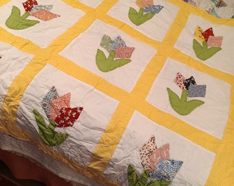 May include: A yellow and white patchwork quilt with a floral design. The quilt features a repeating pattern of tulips made from different fabric scraps. The quilt is made with a variety of fabrics, including floral prints, polka dots, and solid colors.