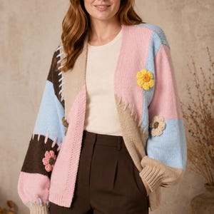 May include: A colour-blocked knit cardigan featuring panels of light pink, baby blue, dark brown, and beige. The garment is accented with three-dimensional crocheted flower appliqués in yellow and beige, and decorative whip-stitch detailing.