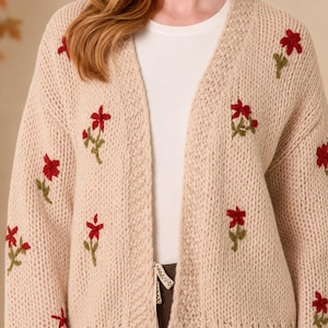 May include: A beige open-front knit cardigan featuring long sleeves and a ribbed hem. The garment is decorated with embroidered red flowers with green stems scattered across the front and sleeves. It is worn over a plain white t-shirt.