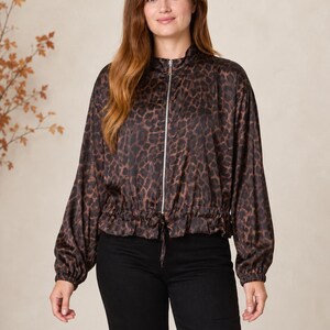 Puede incluir: Una chaqueta de manga larga con estampado de leopardo en tonos marrón y negro, cierre frontal de cremallera plateada, cuello alto y dobladillo elástico con cordón ajustable, combinada con pantalones negros lisos.