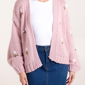 May include: A chunky knit cardigan in a soft dusty pink hue, featuring an open front design and long sleeves. The garment is decorated with small, embroidered white floral motifs with green stems scattered across the front and sleeves.