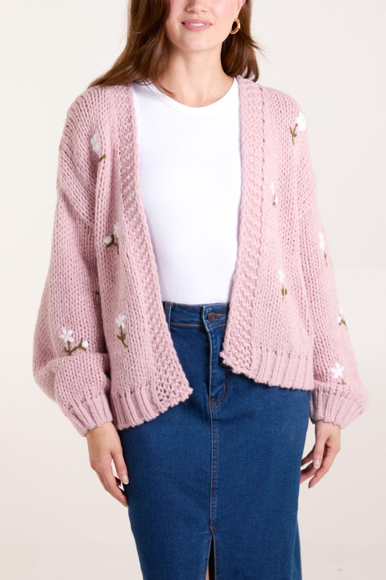 May include: A chunky knit cardigan in a soft dusty pink hue, featuring an open front design and long sleeves. The garment is decorated with small, embroidered white floral motifs with green stems scattered across the front and sleeves.