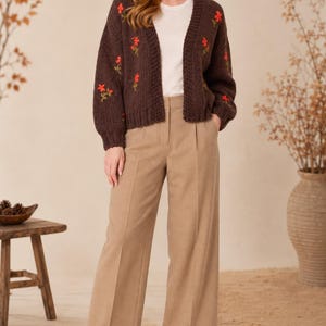 May include: A dark brown knit cardigan featuring embroidered red floral accents, worn over a plain white t-shirt. The outfit is completed with wide-leg tan trousers and dark brown ankle boots, creating a coordinated ensemble.