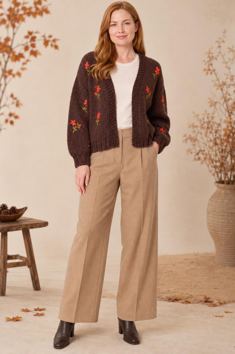 May include: A dark brown knit cardigan featuring embroidered red floral accents, worn over a plain white t-shirt. The outfit is completed with wide-leg tan trousers and dark brown ankle boots, creating a coordinated ensemble.