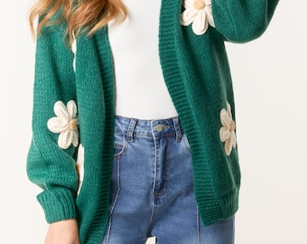 May include: A dark green knit cardigan featuring long sleeves and an open front design. The garment is decorated with large, three-dimensional white flower appliqués that have tan centers, distributed across the front and sleeves of the sweater.