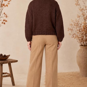 May include: A dark brown long-sleeved knit sweater with a ribbed crew neckline, cuffs, and hem, paired with tan wide-leg trousers featuring a pressed crease down the center of each leg and black block-heeled ankle boots.