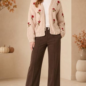 May include: A beige open-front knit cardigan featuring embroidered red floral patterns on the sleeves and front panels, worn over a white top with dark brown wide-leg trousers that have a center seam detail and beige ankle boots.