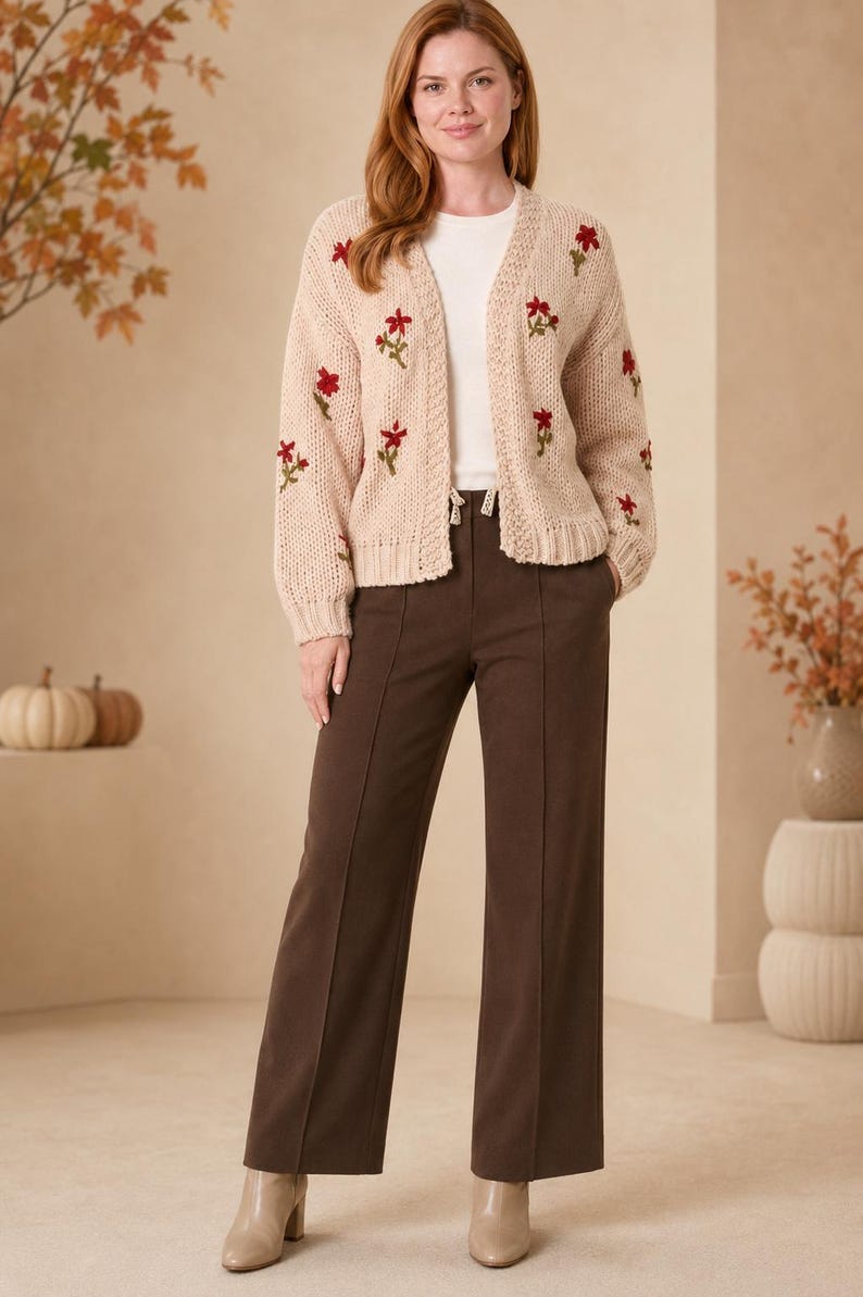 May include: A beige open-front knit cardigan featuring embroidered red floral patterns on the sleeves and front panels, worn over a white top with dark brown wide-leg trousers that have a center seam detail and beige ankle boots.