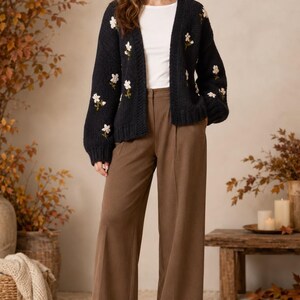 May include: A black open-front knit cardigan featuring embroidered white floral patterns on the sleeves and front, worn over a plain white top with brown wide-leg trousers and black ankle boots.