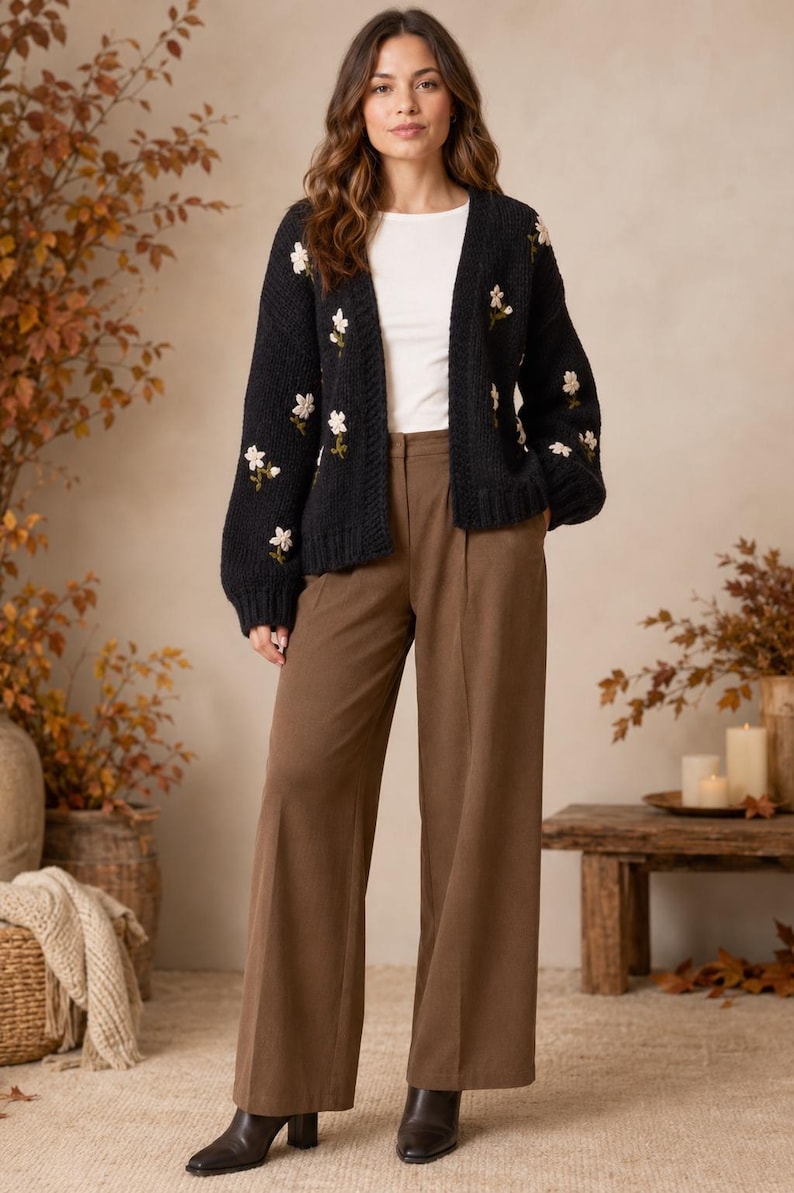 May include: A black open-front knit cardigan featuring embroidered white floral patterns on the sleeves and front, worn over a plain white top with brown wide-leg trousers and black ankle boots.