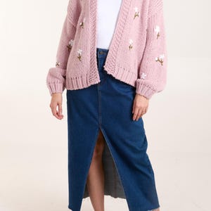 May include: A chunky knit cardigan in a soft dusty pink hue featuring embroidered white floral accents with green stems. The cardigan is worn over a plain white t-shirt and a long, dark blue denim skirt with a front slit, paired with white sneakers.