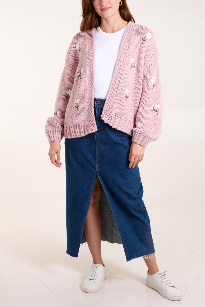 May include: A chunky knit cardigan in a soft dusty pink hue featuring embroidered white floral accents with green stems. The cardigan is worn over a plain white t-shirt and a long, dark blue denim skirt with a front slit, paired with white sneakers.
