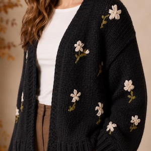 May include: A black knit cardigan featuring an open front, long sleeves, and ribbed cuffs. The garment is decorated with embroidered white flowers with green stems and leaves scattered across the front panels and the outer side of the sleeves.