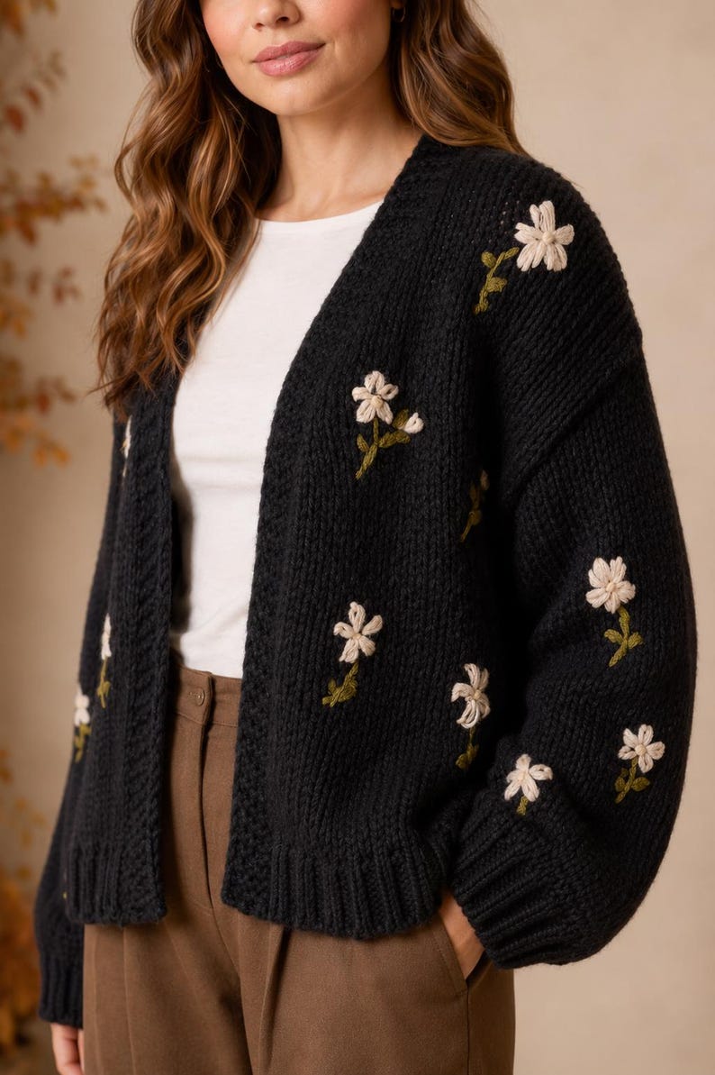 May include: A black knit cardigan featuring an open front, long sleeves, and ribbed cuffs. The garment is decorated with embroidered white flowers with green stems and leaves scattered across the front panels and the outer side of the sleeves.