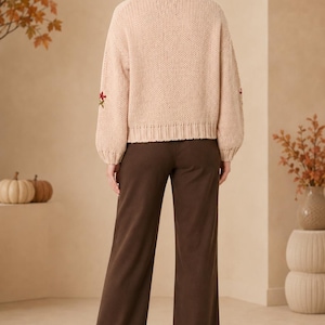 May include: A beige long-sleeve knit sweater with ribbed cuffs and hem, featuring small embroidered floral accents on the sleeves, paired with solid dark brown wide-leg trousers and beige block-heeled ankle boots.