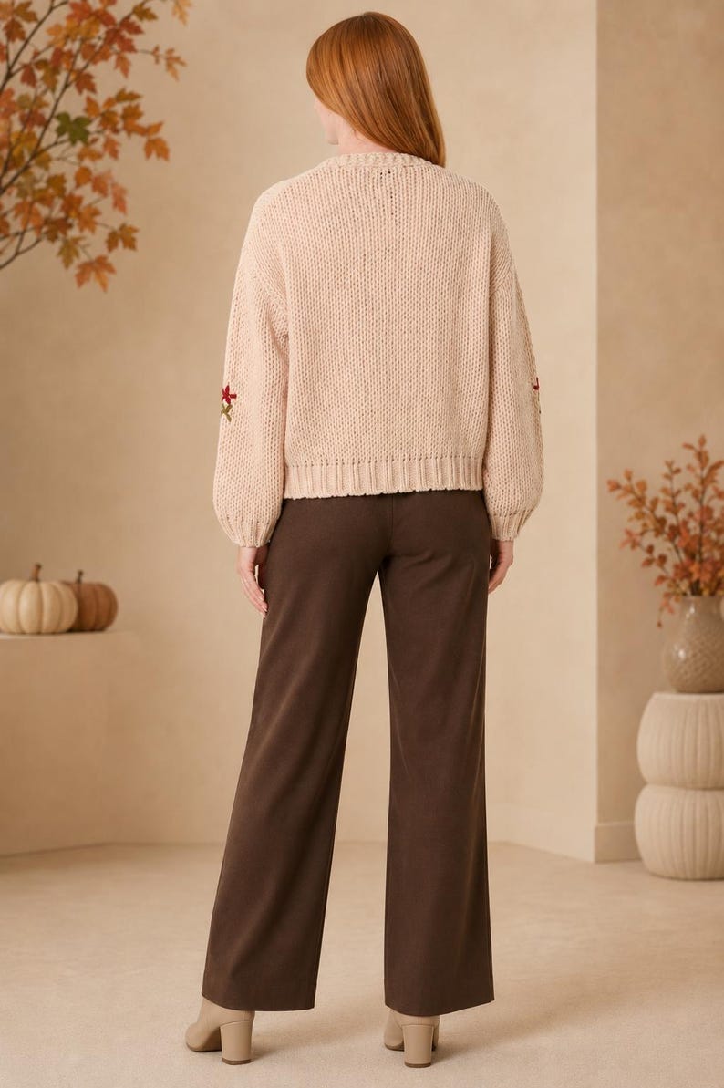 May include: A beige long-sleeve knit sweater with ribbed cuffs and hem, featuring small embroidered floral accents on the sleeves, paired with solid dark brown wide-leg trousers and beige block-heeled ankle boots.