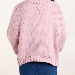 May include: A light pink, chunky knit sweater with long sleeves and a ribbed hemline, paired with a dark blue denim skirt. The sweater features a thick, textured yarn construction and a crew neckline, worn over a simple blue denim garment.