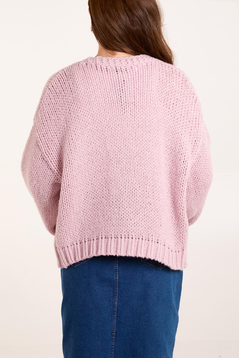 May include: A light pink, chunky knit sweater with long sleeves and a ribbed hemline, paired with a dark blue denim skirt. The sweater features a thick, textured yarn construction and a crew neckline, worn over a simple blue denim garment.