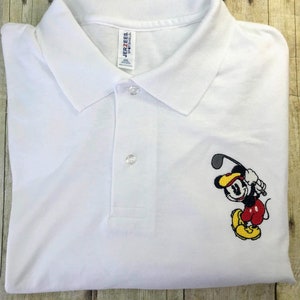Puede incluir: Camiseta polo blanca con una imagen bordada de Mickey Mouse jugando al golf. La camiseta tiene una etiqueta de la marca Jerzees.