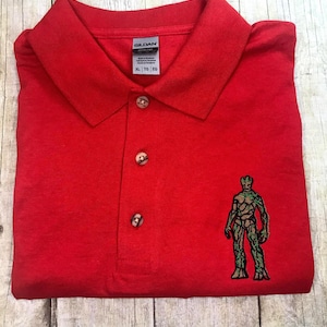 Può includere: Polo rosso con un'immagine ricamata verde di una figura umanoide simile a un albero. La polo ha un colletto abbottonato e un'etichetta del marchio Gildan con la taglia XL.