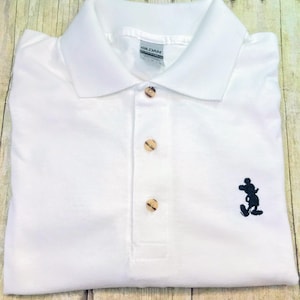 Puede incluir: Polo blanco con una silueta de Mickey Mouse bordada en negro en el lado derecho. La camisa tiene un cuello abotonado y botones marrones.