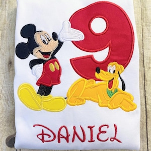 Puede incluir: Camiseta blanca con bordado de Mickey Mouse, Pluto y el número 9 en rojo. Mickey viste de rojo y amarillo, Pluto es amarillo, y el nombre "DANIEL" está bordado en rojo. Ropa infantil.