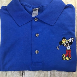 Könnte beinhalten: Ein königsblaues Poloshirt mit einem gestickten Bild von Mickey Mouse beim Golfspielen. Mickey trägt ein rotes Hemd, einen gelben Hut und schwarze Schuhe. Er hält einen Golfschläger in der Hand und befindet sich in einer Golfhaltung.