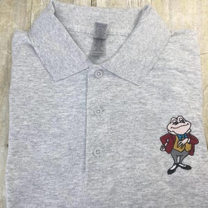 Könnte beinhalten: Ein graues Poloshirt mit einem gestickten Bild eines Frosches, der eine rote Weste und einen Zylinder trägt. Der Frosch steht mit verschränkten Armen da und hat ein Lächeln im Gesicht.