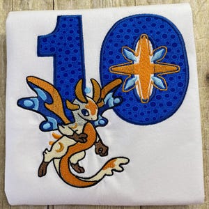 May include: White t-shirt with a blue and orange embroidered design. The design features the number 10, a dragon, and a star.