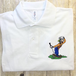 May include: White polo shirt with an embroidered image of Goofy playing golf. Goofy is wearing a green hat, orange shirt, blue pants, and brown shoes. He is holding a golf club and looking to the right.
