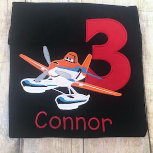 May include: Black t-shirt with a red number 3, an orange and white airplane, and the name Connor in red embroidery.