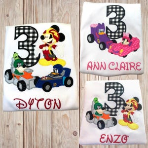 May include: Three personalized white t-shirts with a black and white checkered number 3, a Mickey Mouse character, and a race car design. The shirts are personalized with the names "Dayton", "Ann Claire", and "Enzo".
