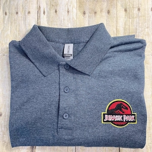Può includere: Una polo grigia scura con un logo Jurassic Park ricamato in giallo, rosso e nero sul lato destro del petto.