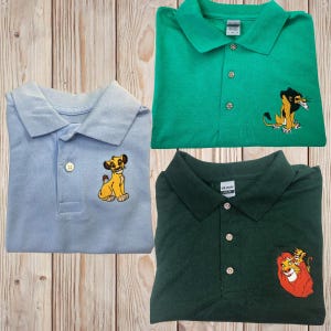 May include: Three polo shirts with embroidered lion king characters. The first shirt is light blue with a yellow lion cub. The second shirt is green with a yellow lion. The third shirt is dark green with a yellow lion and a yellow lion cub.