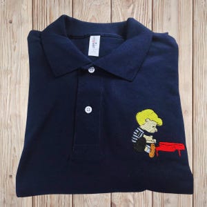 Può includere: Polo blu navy con colletto bianco abbottonato e un piccolo personaggio dei cartoni animati ricamato sul davanti. Il personaggio ha i capelli gialli, una maglietta a righe ed è seduto su una panchina rossa.